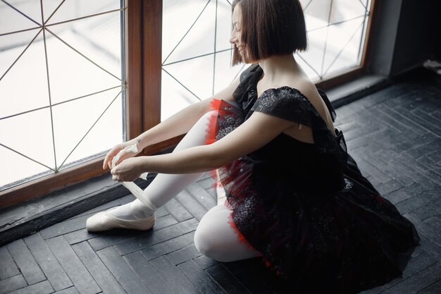 Bailarina de ballet con tutú negro y zapatillas de punta sentada cerca de una ventana grande