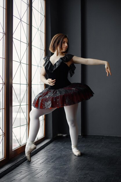 Bailarina de ballet en tutú negro y zapatos puntiagudos de pie cerca de una ventana grande