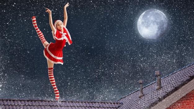Bailarina de garota dançando com um presente nas mãos em uma fantasia de Papai Noel no telhado de uma casa na noite de Natal contra o fundo do céu estrelado