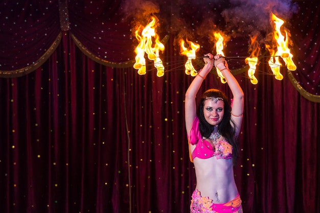 Bailarina de fuego exótica con traje rosa brillante Aparato llameante giratorio sobre la cabeza en el escenario con cortina roja