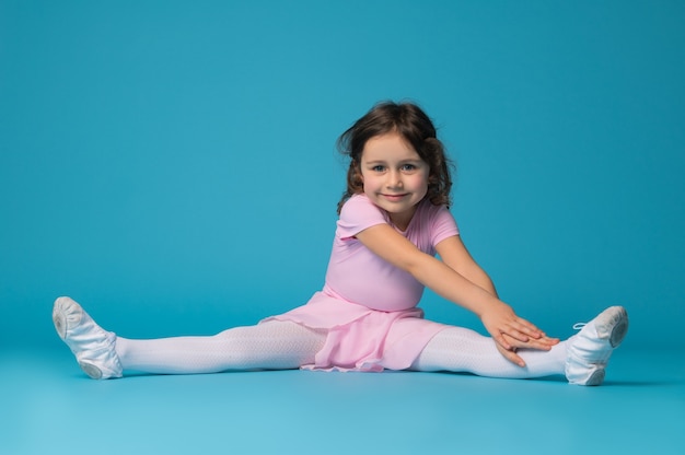 Bailarina hermosa niña en vestido rosa sentada sobre un fondo azul, practicando y estirando su cuerpo