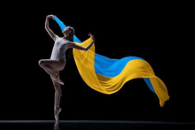 Bailarina jovem dançando com pano pintado em cores azuis e amarelas da bandeira da Ucrânia no fundo escuro do estúdio