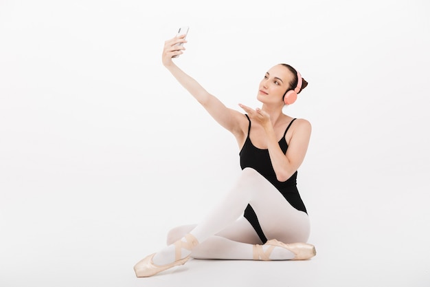 Bailarina joven caucásica en auriculares tomando foto selfie en teléfono móvil aislado sobre fondo de pared blanca