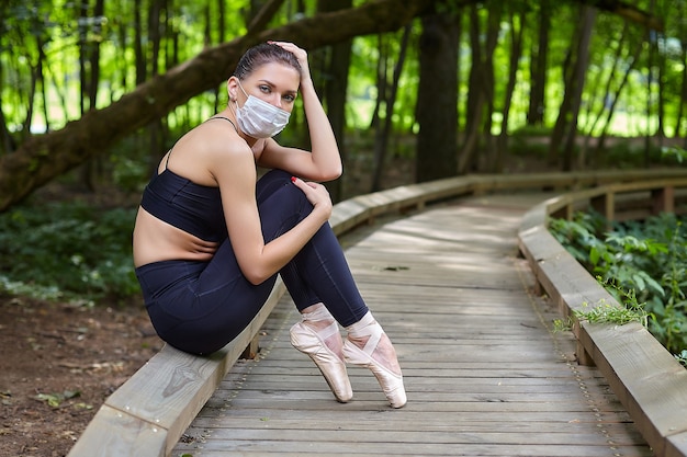 Una bailarina con máscara y zapatos de punta está haciendo ejercicios en un camino de madera en un parque forestal. concepto de entrenamiento al aire libre seguro