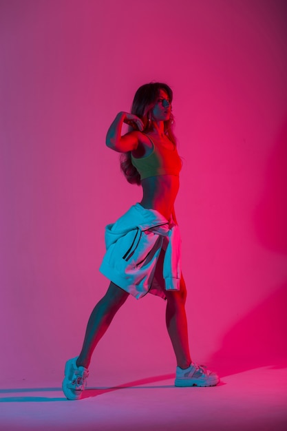Bailarina de mujer joven de moda en una ropa juvenil de moda en zapatillas con estilo bailando en el estudio con color neón rosa brillante en estilo discoteca. Chica disfruta de un baile en el estudio con luz multicolor.