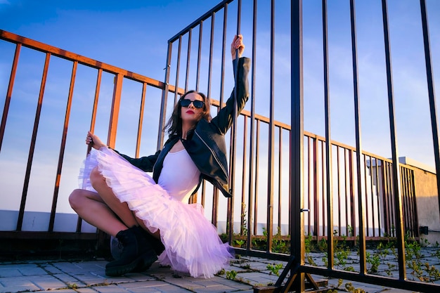 Bailarina en tutú sentada en la valla en el techo xAat atardecer con botas y una chaqueta