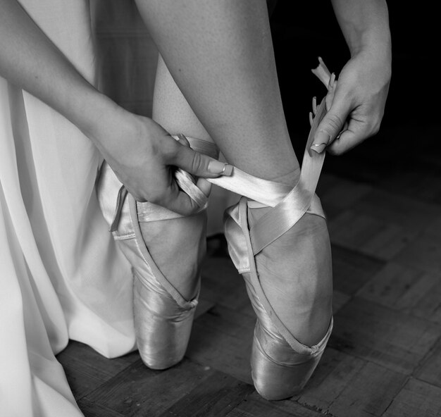 Foto una bailarina con un vestido blanco y zapatos de punta se sienta y se pone zapatos de pointe sobre un fondo negro