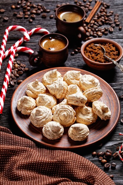 Baiser-Kekse serviert auf einem Teller mit Weihnachtszuckerrohr, Kaffeetassen, Kaffeebohnen, braunem Zucker und Zimtstangen auf dunklem Holzhintergrund, Draufsicht, Nahaufnahme