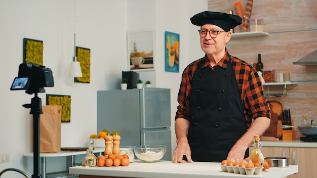 Baker grabando para podcast en la cocina mientras tamiza la harina en la mesa. Influencer chef blogger jubilado que usa tecnología de Internet para comunicarse, disparar, bloguear en las redes sociales con equipos digitales