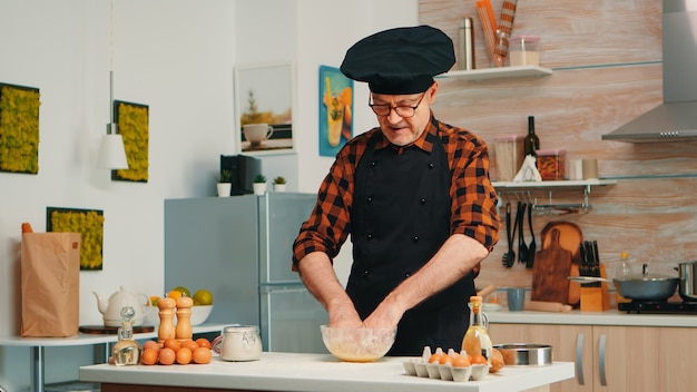 Baker kending masa en la mesa de la cocina vistiendo delantal y bonete. Cocinero anciano jubilado con rociado uniforme, tamizado tamizando ingredientes crudos horneando a mano pizza casera, pan.