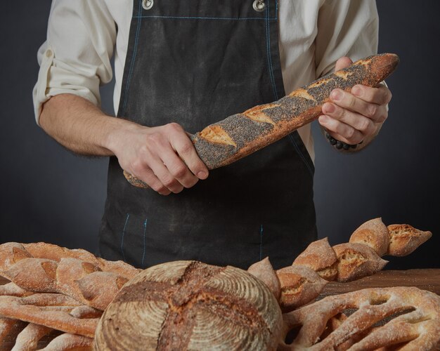 Baker segura uma baguete com sementes de papoula em um fundo escuro e uma variedade de pães na mesa