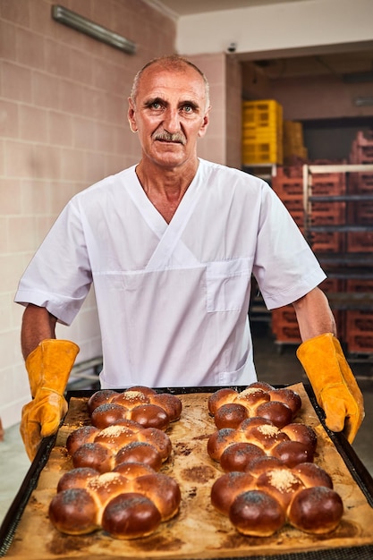 Baker segura uma bandeja com pão quente fresco nas mãos contra o fundo das prateleiras com pastelaria na padaria Produção industrial de pão