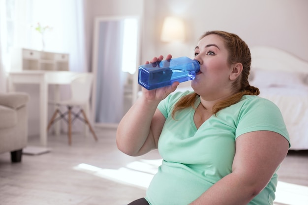 Balance de agua. Mujer joven con sobrepeso sentada en la estera de yoga mientras bebe agua de la botella