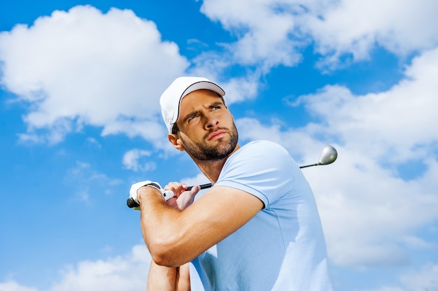 Balanço profissional. Vista de baixo ângulo de um jogador de golfe jovem e confiante balançando seu driver e olhando para longe com o céu azul como pano de fundo