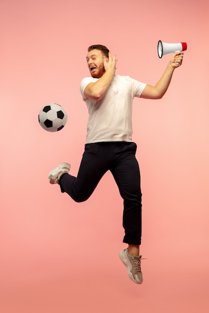 Ball-Fußball-Fan fangen. Ganzaufnahme des jungen hochspringenden Mannes einzeln auf rosa Studiohintergrund. Männliches kaukasisches Modell. Exemplar. Menschliche Emotionen, Gesichtsausdruck, Sportkonzept.