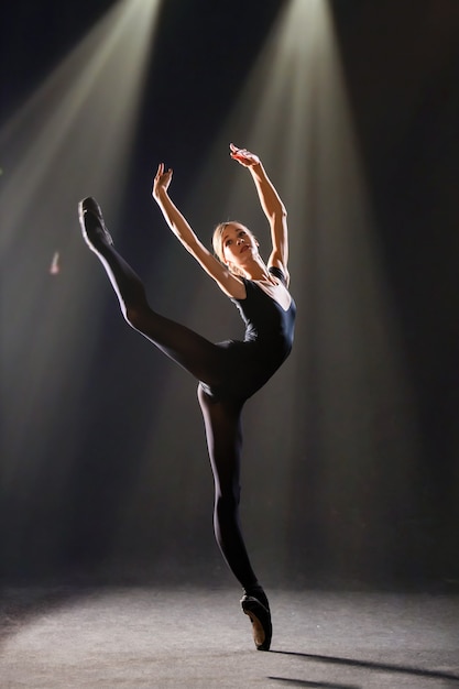 Ballerina im eng anliegenden Anzug tanzt auf schwarzem Hintergrund auf Spitzenschuhen, Silhouette wird von Farbquellen beleuchtet.