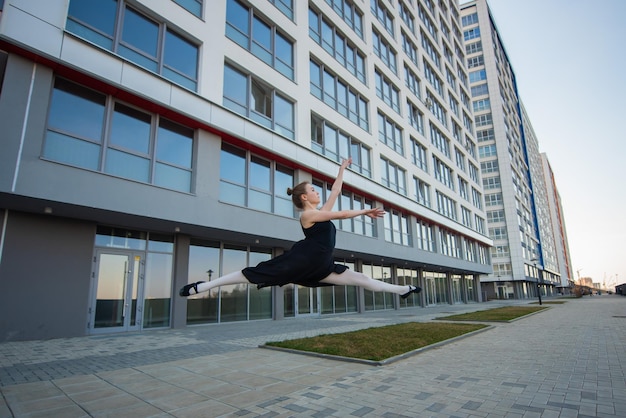 Ballerina im Tutu posiert vor einem Wohnhaus Schöne junge Frau in schwarzem Kleid und Spitzenschuhen springt mit unglaublicher Flexibilität Ballerina führt elegantes Springen mit Durchbiegung durch