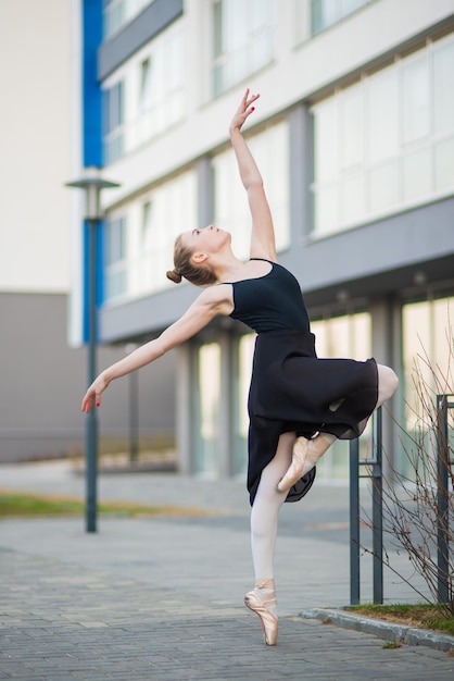 Ballerina in einem Tutu posiert vor dem Hintergrund eines Wohnhauses Schöne junge Frau in schwarzem Kleid und Spitzenschuhen, die draußen Ballett tanzen