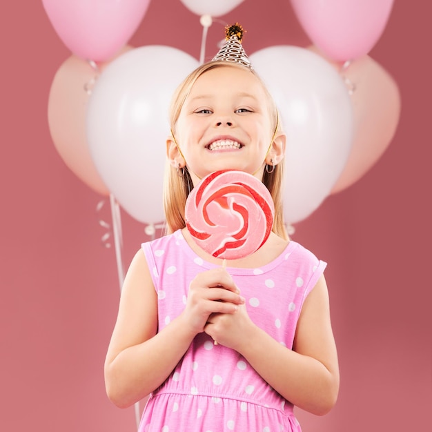 Balões, pirulito e retrato de menina em fundo rosa para celebração de festa de aniversário e dia especial Feliz, emocionado e jovem criança bonita sorri com doces doces e sobremesa no estúdio