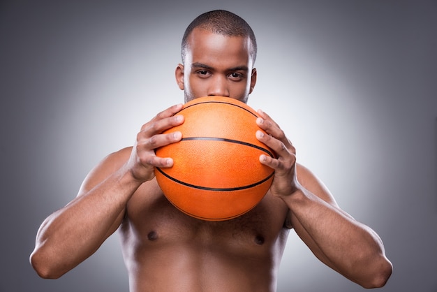 El baloncesto es mi vida. Joven africano descamisado que oculta parte de su rostro detrás de una pelota de baloncesto