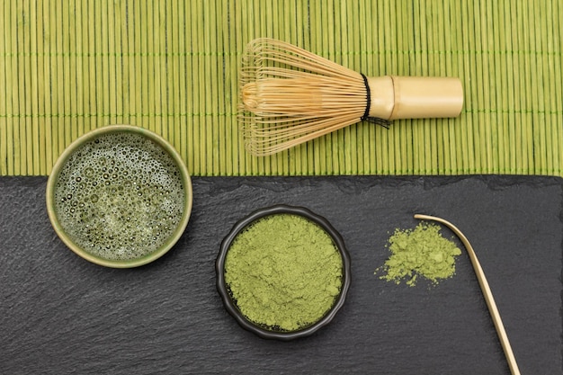 Bambusbesen auf grüner Serviette aus Holzstäbchen Matcha-Teepulver und Matcha-Tee auf Steinbrett flach legen
