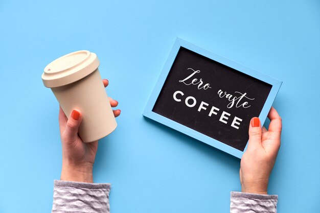 Bambuskaffeetasse, halten Tasse oder Reisebecher in weiblicher Hand auf blauem Minzhintergrund. Kreative flache Lage, Bild, Text "Zero Waste Coffee" auf Tafel. Umweltfreundlicher Reisebecher ohne Abfall.