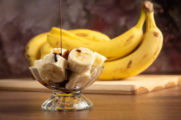 Banana cortada em fatias em uma jarra de vidro com cobertura de chocolate e mais bananas ao fundo, fundo abstrato escuro, foco seletivo.