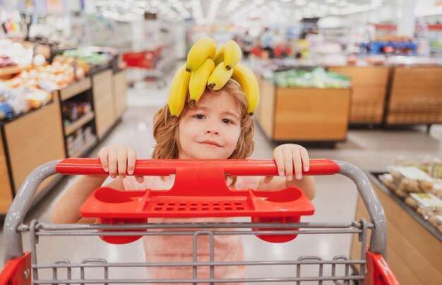 Banana na cabeça de criança engraçada. Garoto engraçado com carrinho de compras comprando comida na mercearia ou supermercado. Rosto de crianças surpresas.