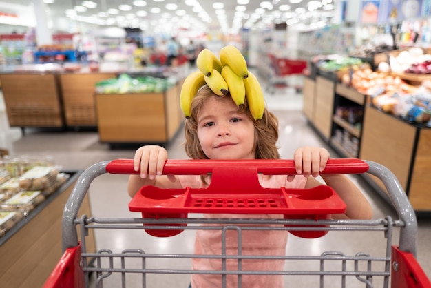 Banana na cabeça de criança engraçada. Garoto engraçado com carrinho de compras, comprar comida no supermercado ou supermercado. Rosto de crianças surpresas.