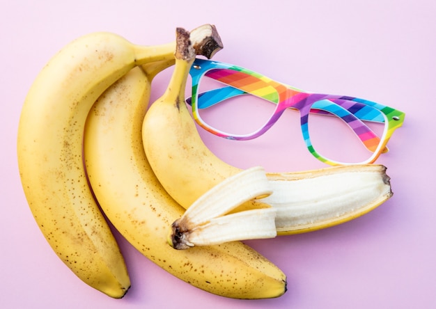 Bananas amarelas e óculos de cores LGBT em fundo rosa claro. Acima vista