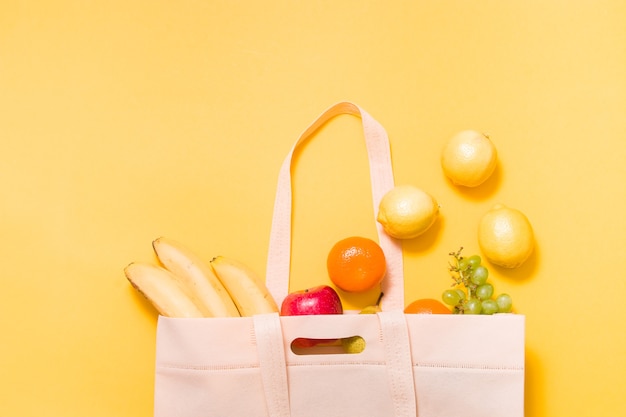 Bananen, Mandarinen, Trauben, Apfel, Birne und Zitronen in einer Stoff-Einkaufstasche auf gelber Oberfläche