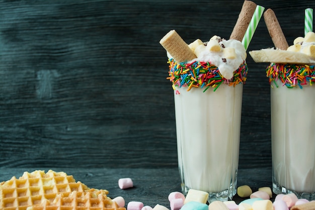 Bananenmilchshake mit Eiscreme und Schlagsahne, Marshmallows, Keksen, Waffeln, serviert in einer Glasschale mit Süßwarendressing. Dunkler hölzerner Hintergrund.