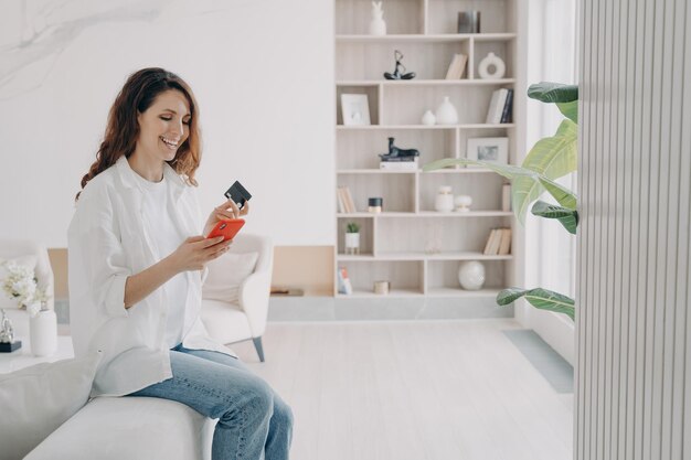 Banca en línea Feliz mujer sosteniendo un teléfono inteligente con tarjeta de crédito bancaria hace pagos sin efectivo en casa