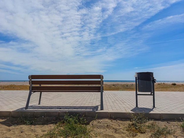 Un banco y un bote de basura están en una acera cerca de una playa.