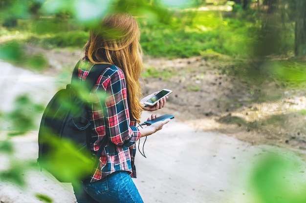 Banco de potência e smartphone nas mãos de uma garota ruiva em uma camisa em uma gaiola com uma mochila preta, no fundo de uma estrada na floresta.