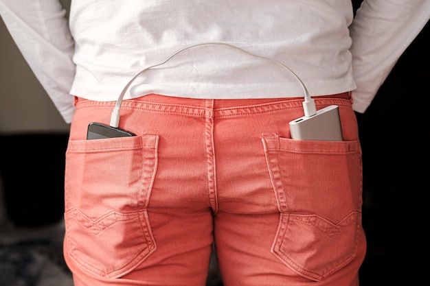 Foto el banco de energía se encuentra en el bolsillo trasero de los jeans de color coral y en otro hay un teléfono móvil que se está cargando