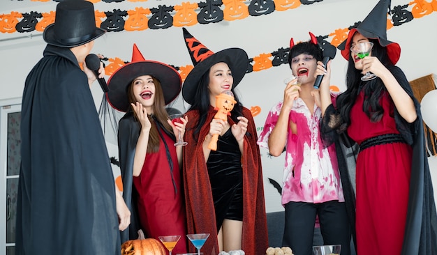 Bande junger Asiaten in Kostümhexe, Zauberer mit Halloween-Party zum Singen eines Liedes und Trinkens, Dessert im Zimmer. Gruppe jugendlich Thai mit Halloween feiern. Konzeptparty Halloween zu Hause.