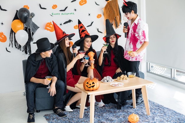 Foto bande junger asiaten in kostümhexe, zauberer mit halloween-party zum singen eines liedes und trinkens, dessert im zimmer. gruppe jugendlich thai mit halloween feiern. konzeptparty halloween zu hause.