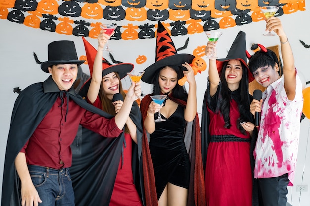 Bande junger Asiaten in Kostümhexe, Zauberer mit Halloween-Party zum Singen eines Liedes und Trinkens, Dessert im Zimmer. Gruppe jugendlich Thai mit Halloween feiern. Konzeptparty Halloween zu Hause.