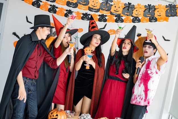 Bande junger Asiaten in Kostümhexe, Zauberer mit Halloween-Party zum Singen eines Liedes und Trinkens, Dessert im Zimmer. Gruppe jugendlich Thai mit Halloween feiern. Konzeptparty Halloween zu Hause.