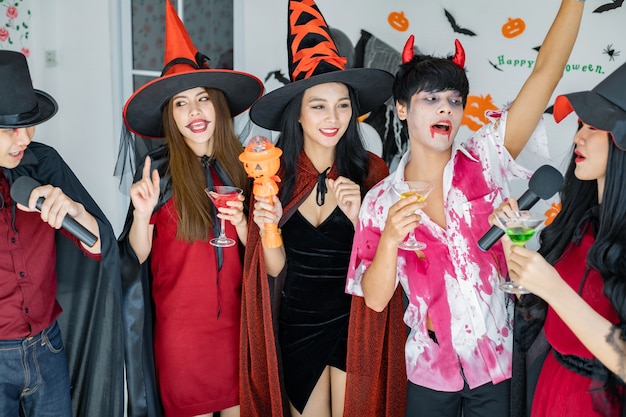 Foto bande junger asiaten in kostümhexe, zauberer mit halloween-party zum singen eines liedes und trinkens, dessert im zimmer. gruppe jugendlich thai mit halloween feiern. konzeptparty halloween zu hause.