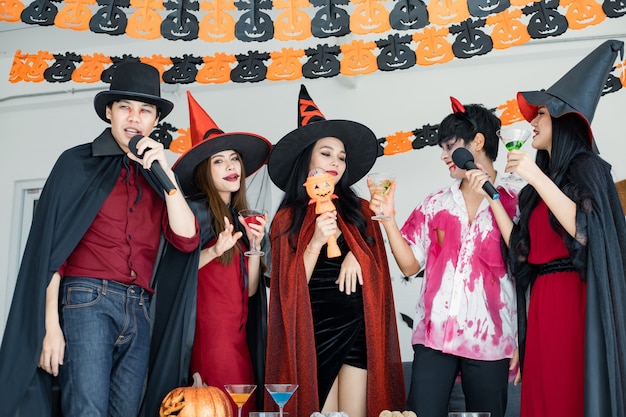 Bande junger Asiaten in Kostümhexe, Zauberer mit Halloween-Party zum Singen eines Liedes und Trinkens, Dessert im Zimmer. Gruppe jugendlich Thai mit Halloween feiern. Konzeptparty Halloween zu Hause.