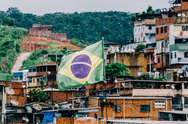 Foto bandeira brasileira vibrante ondulando em primeiro plano de uma favela no brasil