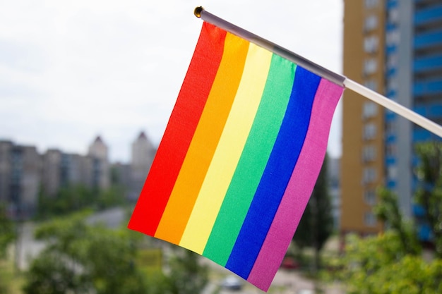 Bandeira LGBT do arco-íris contra o céu azul, árvores verdes, ruas da cidade e prédios de vários andares