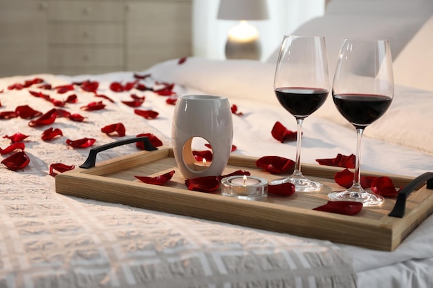 Bandeja con copas de vino tinto, velas y pétalos de rosa en la cama en la habitación