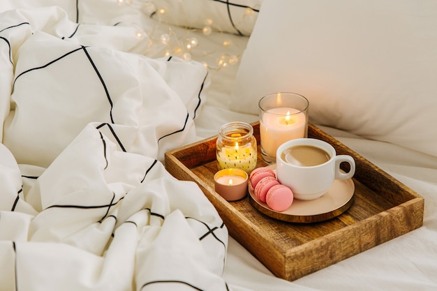 Bandeja de madeira com café e velas na cama. lençóis brancos com cobertor listrado e travesseiro. café da manhã na cama. conceito de hygge.