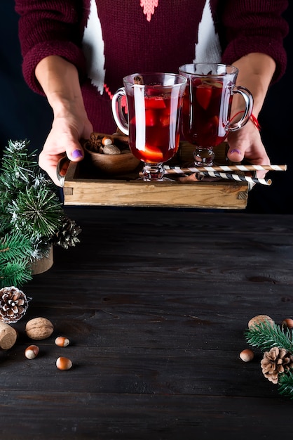 Bandeja de madeira com vinho quente caseiro quente com frutas e especiarias nas mãos femininas em um escuro