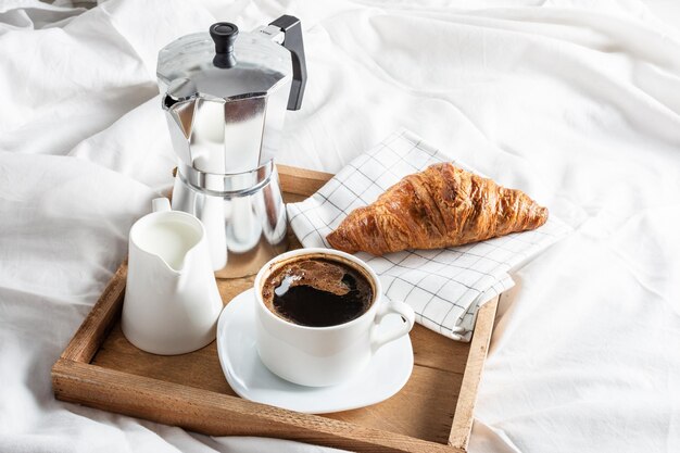 Bandeja de madeira com xícara de café preto jarro de leite e croissant na cama de linho branco