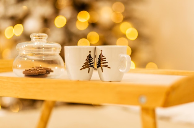 Una bandeja de madera se encuentra sobre la cama con el telón de fondo de las luces navideñas. En la bandeja hay un tarro de galletas y dos tazas.