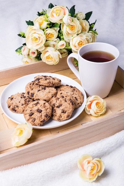 Bandeja de madera con galletas de chocolate, taza de té y flores sobre fondo blanco.